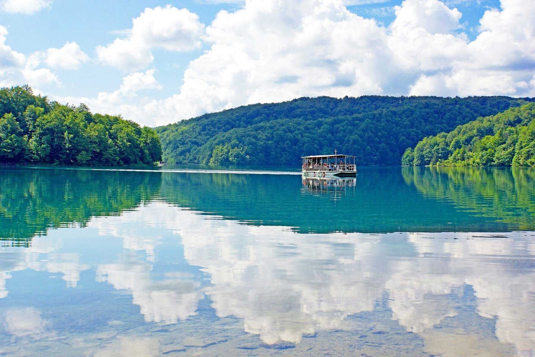 Un bateau électrique chargé de visiteurs traverse le lac Kozjak, entre les collines boisées qui s’y reflètent