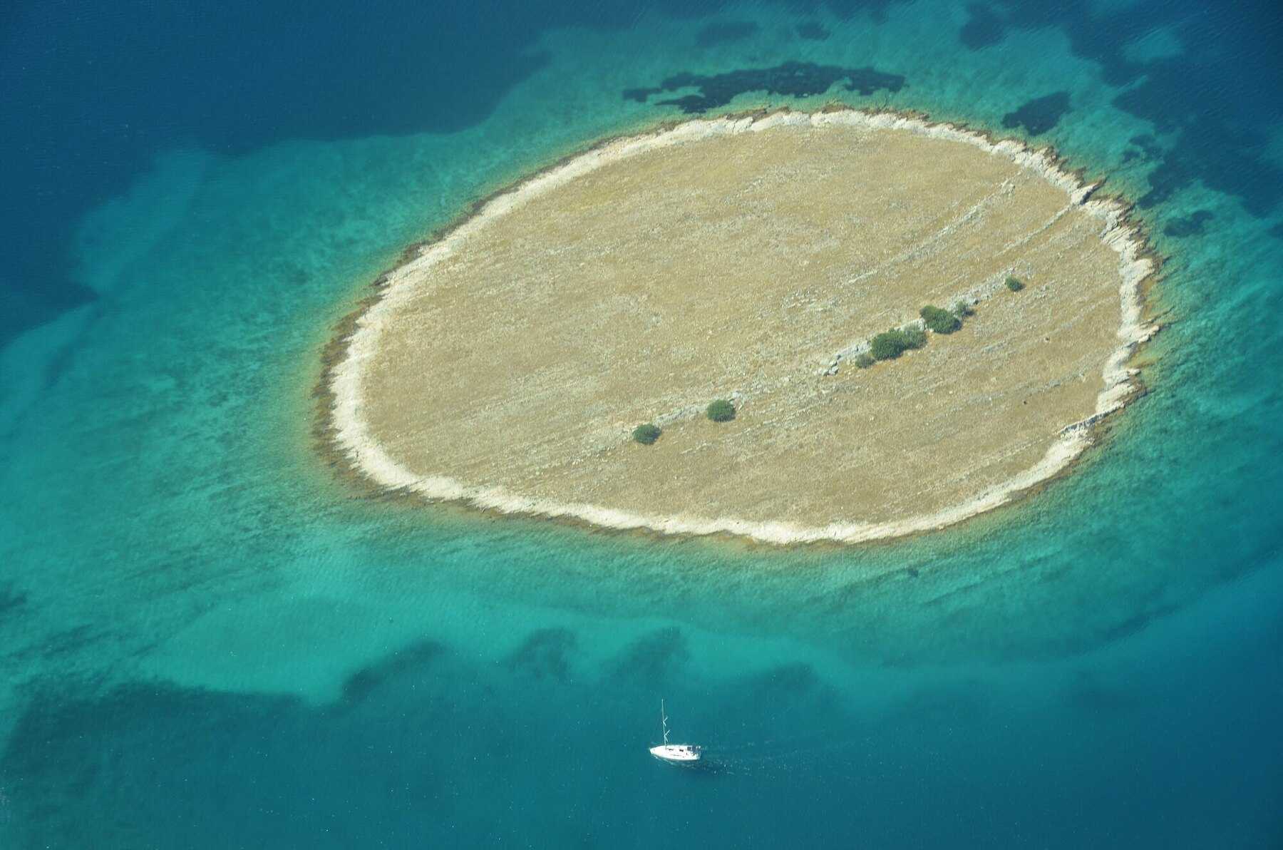 Un îlot nu entouré d'eau turquoise et un voilier, vus d'avion