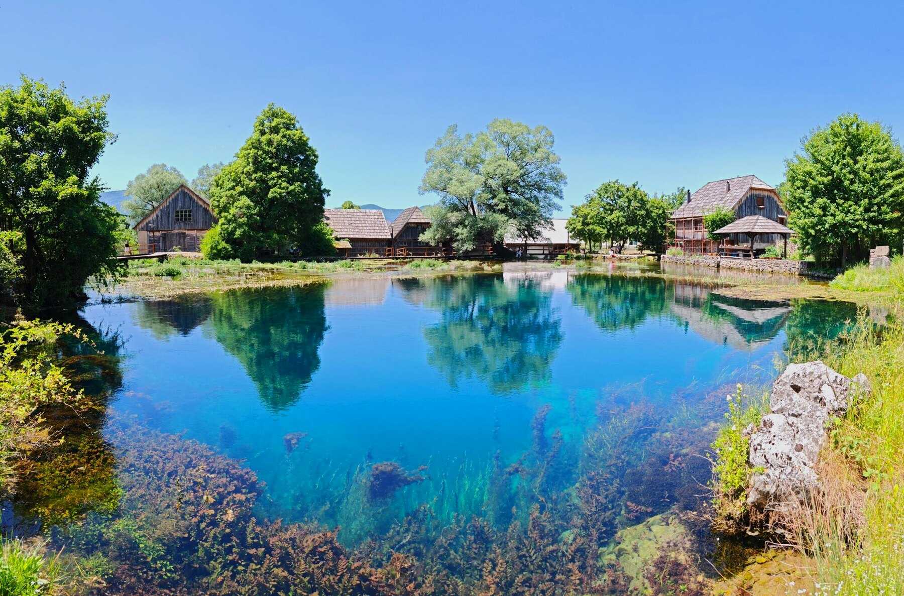 Une source de la Gacka d'un bleu profond, bordée de maisons et d'arbres