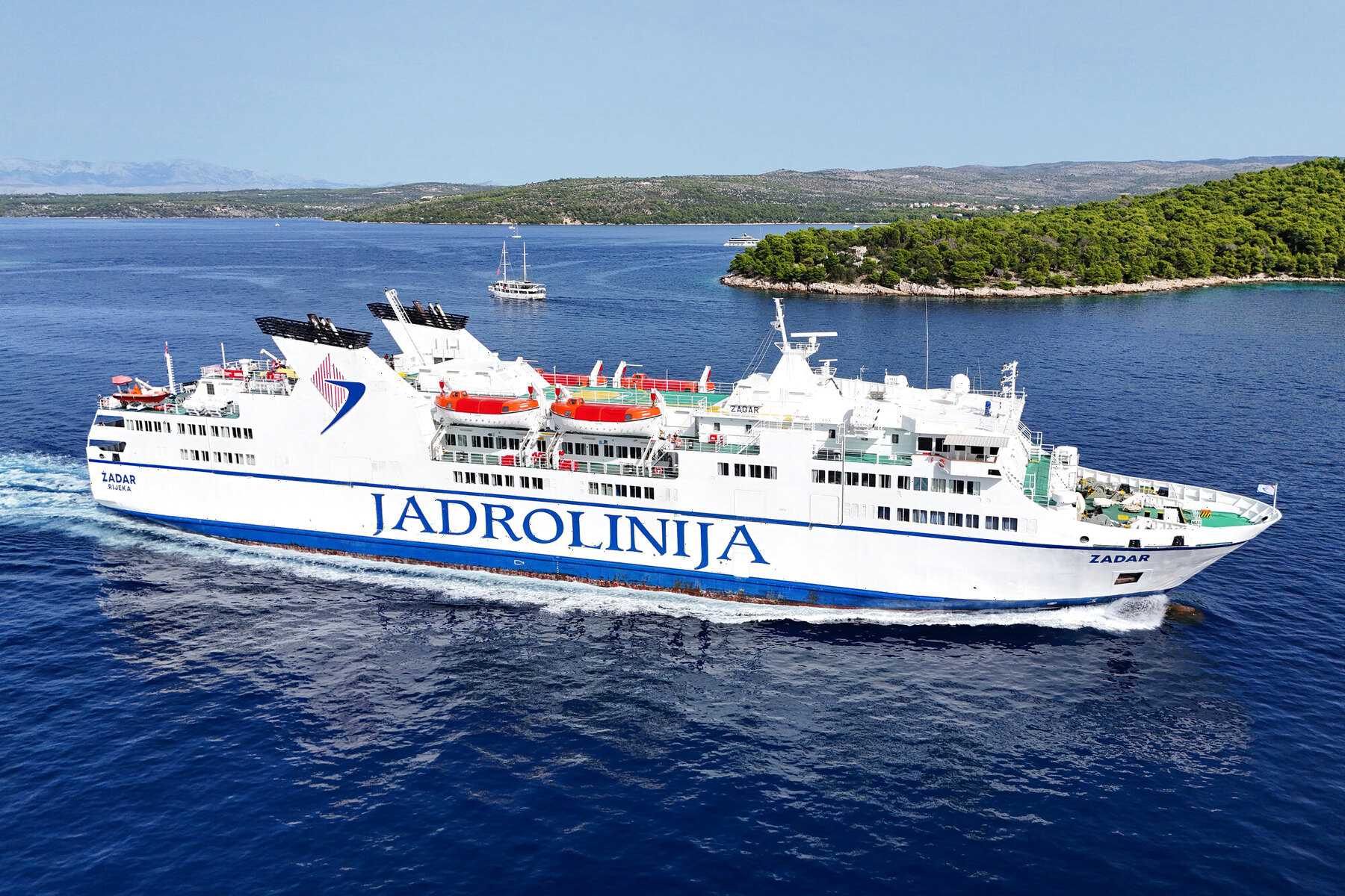 Le car-ferry Jadrolinija Zadar en pleine mer, sous le soleil, avec une île boisée à l'horizon