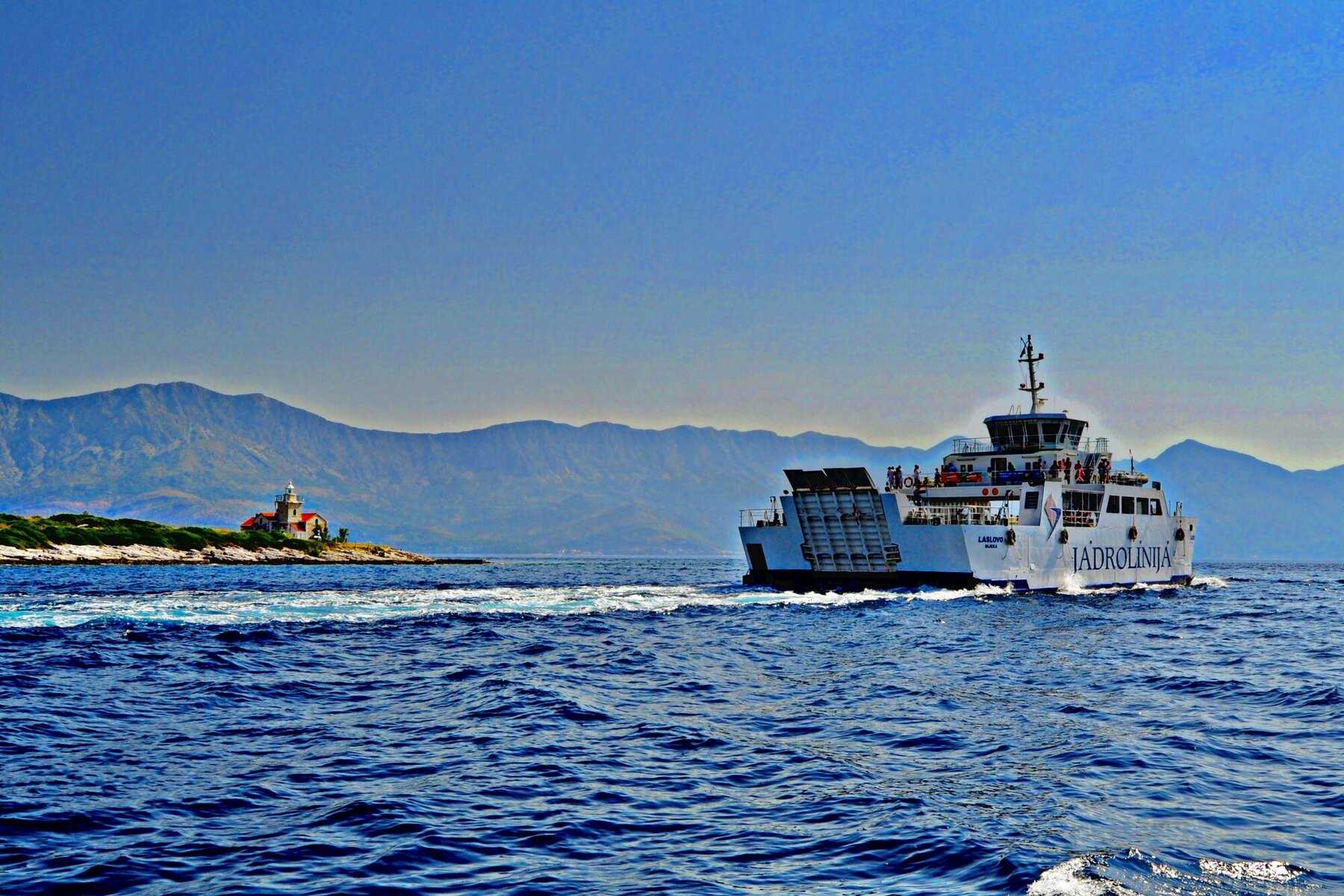 Un car-ferry Jadrolinija en pleine mer, au large d'un petit phare, avec les montagnes de la côte dalmate à l'horizon